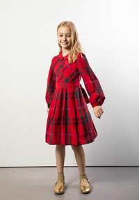 Czerwona sukienka w kratę z długimi rękawami, kołnierzykiem i wiązaniem w talii. Sukienka ma plisowane falbany u dołu, noszona z złotymi butami i beżowymi skarpetkami.