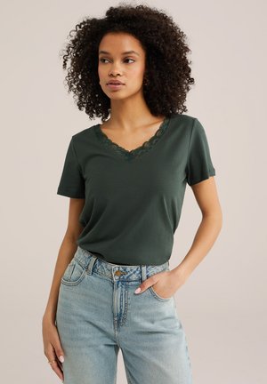 Žena s kučeravými vlasmi, ktorá má na sebe tmavozelený top s krátkym rukávom a výstrihom do V lemovaný čipkou, zastrčený do svetlomodrých džínsov s vysokým pásom, stojaca s jednou rukou v vrecku.