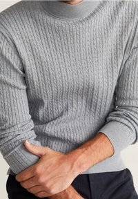 Suéter de punto gris claro con textura acanalada, cuello alto redondeado y mangas ajustadas. Vista en primer plano de manos sobre la tela.