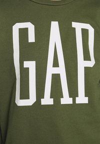 Olivgrön sweatshirt med stora vita blockbokstäver som stavar "GAP" på framsidan. Tillverkad av mjukt, texturerat tyg med klassisk rund hals.