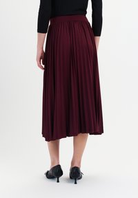 Frau, die einen bordeauxfarbenen plissierten Midikleid trägt und schwarze Kitten-Heels Schuhe, die vor einem weißen Hintergrund steht, von hinten betrachtet.