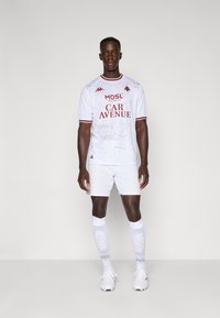 Weißes Fußballtrikot mit roten Akzenten, ausgestattet mit Markenlogos und einem grafischen Design. Kombiniert mit weißen Shorts und knielangen weißen Socken.
