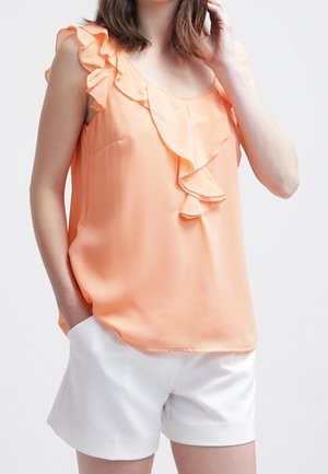 Blusa senza maniche color pesca con dettagli in raso lungo il colletto; tessuto morbido e leggero. Abbinata a pantaloni corti bianchi.