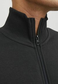 Veste noire à col montant en tissu doux avec une texture côtelée. Dispose d'une fermeture éclair complète avec un curseur lisse et des coutures plates.