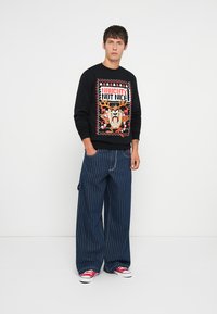 Sudadera negra con un diseño gráfico y el texto "Travieso, no agradable", combinada con pantalones de pinzas azul de pierna ancha y zapatillas rojas.