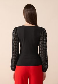 Haut noir avec un corsage ajusté et de longues manches en dentelle transparente présentant un motif géométrique. Porté avec un pantalon rouge en tissu lisse.