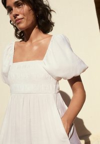 Witte jurk met opgezette mouwen, een vierkante halslijn, een gerimpelde bodice en een vloeiende rok met zijzakken. De gestructureerde stof geeft zachtheid.