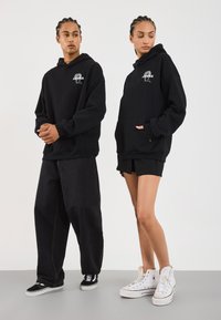 Čierne mikiny s prednými vreckami, zdobené grafickým potlačou. Skombinované s tmavými džínsami a čiernymi teniskami, a čierny krátky outfit s bielymi teniskami.