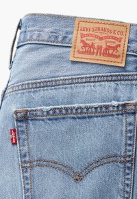 Light blue denim jeans with a red tag, orange stitching, and a tan leather label featuring a logo and text. Back pocket detailing is visible.