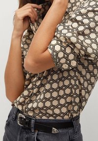 Femme ajustant le col d'une chemise beige avec un motif géométrique hexagonal marron, portant un jean foncé et une ceinture noire avec une boucle argentée.