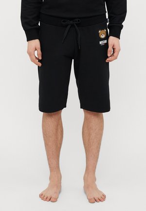 Mustad must reklaamid, millel on elastne vöökoht ja nöör, musta puuvillase kangaga ning vasakul jalal prinditud pruuni karu graafika ja "Moschino" tekst.