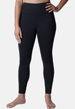 Frau trägt schwarze Leggings mit hoher Taille, steht barfuß mit einer Hand auf der Hüfte und der anderen entspannt seitlich, vor einfachem Hintergrund.