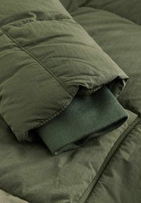 Grøn pufferjakke med ribbet strikket manchet, mat finish og quiltet tekstur. Stoffet er blødt og let krøllet.