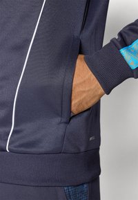 Chaqueta atlética de color navy con ribetes blancos, tela texturizada, puños acanalados y un bolsillo lateral. Presenta detalles en azul con patrones en el puño.