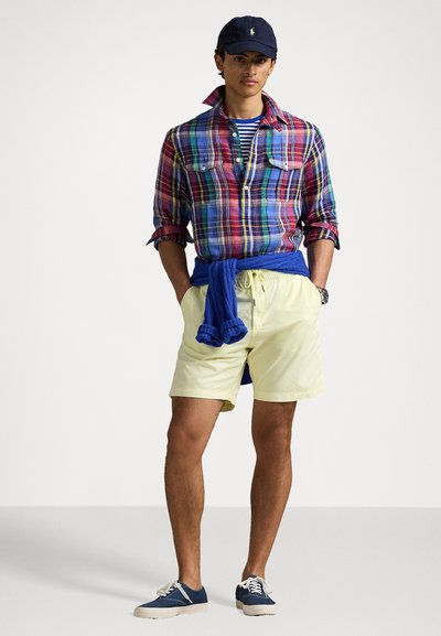 Chemise à carreaux rouge, bleue et verte, portée par-dessus un t-shirt rayé bleu marine et blanc. Un short jaune et des baskets bleues complètent le look.