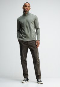 Lichtgrijze zip-up sweater met een kraag, gecombineerd met donkerbruine broeken en zwarte hoge sneakers. De textuur lijkt zacht en gebreid te zijn.