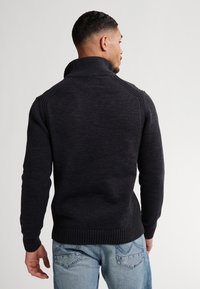Man met zijn rug naar de camera, gekleed in een zwarte gebreide coltrui en lichtblauwe spijkerbroek met zichtbare achterzakken.