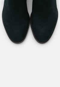 Botas de tobillo de ante verde oscuro con punteras afiladas y una textura suave, con una suela negra cosida en contraste.