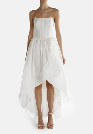 Vestido blanco sin tirantes con un cuerpo ajustado y una falda de capas de largo asimétrico. Tejido suave y ligero; textura lisa, diseño minimalista. Sandalias plateadas.