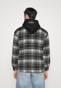 Black and gray plaid flannel jacket with a black hood. Features long sleeves, a relaxed fit, and a tag with "PEGADOR" on the back.