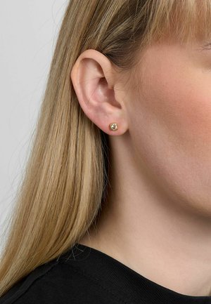 Gros plan sur l'oreille d'une femme aux longs cheveux blonds portant une petite boucle d'oreille carrée en or avec une pierre précieuse claire au centre.