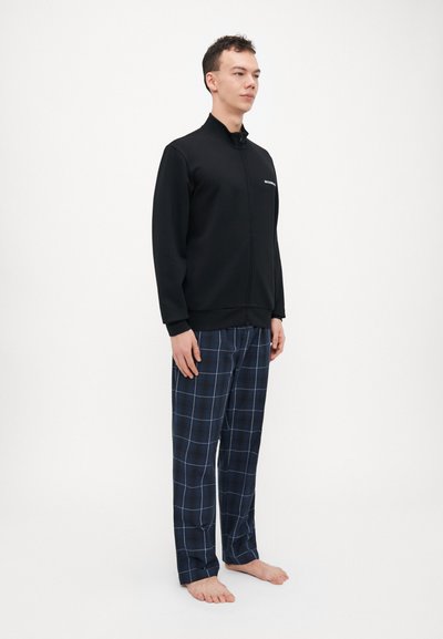 Black zip-up jacket with a high collar, paired with navy plaid pajama pants. Smooth fabric with a subtle sheen, featuring a white logo on the chest.