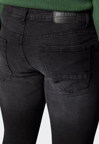 Jeans de mezclilla negra con un diseño ajustado, bolsillos traseros y una etiqueta de cuero con la inscripción "ONLY & SONS". Presentan un leve desgaste y una tela texturizada.