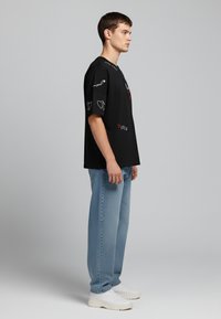 Black oversized t-shirt with printed graphics and text in various colors, paired with light blue straight-leg jeans and white sneakers.