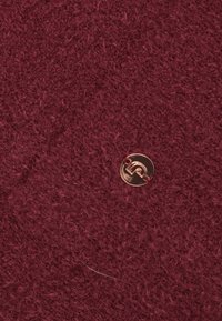 Tissu bordeaux avec une texture douce et duveteuse ; présente un bouton logo rond en or rose bien en vue.