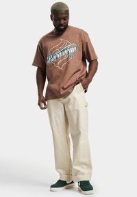 Brown cotton t-shirt with "Rocawear 99" graphic in teal and white, paired with loose cream-colored pants and green sneakers.