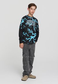 Nastolatek stoi z rękami w kieszeniach, ubrany w czarną koszulę z długim rękawem z niebieskim wzorem błyskawicy, szare dżinsy cargo i beżowe buty.