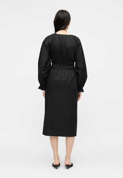 Γυναίκα με μακριά σκούρα μαλλιά, που φοράει μαύρο midi φόρεμα με μακριά μανίκια και ζωνάκι στη μέση και μαύρα mule με τακούνι, στέκεται με την πλάτη στραμμένη.