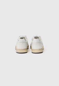 Zapatillas de cuero blanco con detalles en crema, suela de goma y costuras decorativas. Presentan un logotipo en relieve en el talón y la lengüeta.