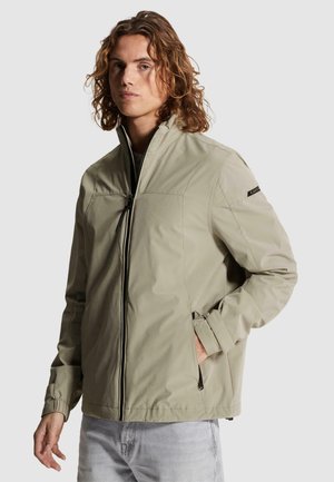 Jeune homme aux cheveux mi-longs portant une veste zippée beige clair avec un col montant et un jean bleu clair, main dans la poche.