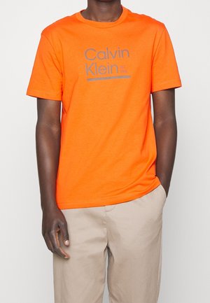 Person iført en knaldrød Calvin Klein T-shirt med gråt logo og beige bukser, stående med den ene hånd i lommen.