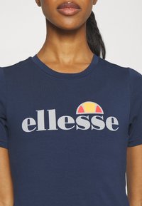 Navy blue T-shirt made of soft fabric with a crew neck, featuring the "ellesse" logo in gray and a colorful sun graphic above it.