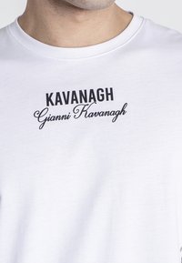 Bílý bavlněný tričko s černým potiskem: "Kavanagh" v tučném písmu, s "Gianni Kavanagh" ve stylu kurzívy pod tím.