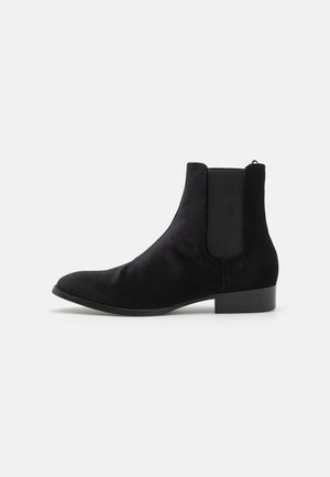 Bottine Chelsea en daim noir avec panneau latéral élastique et petit talon sur fond clair uni.