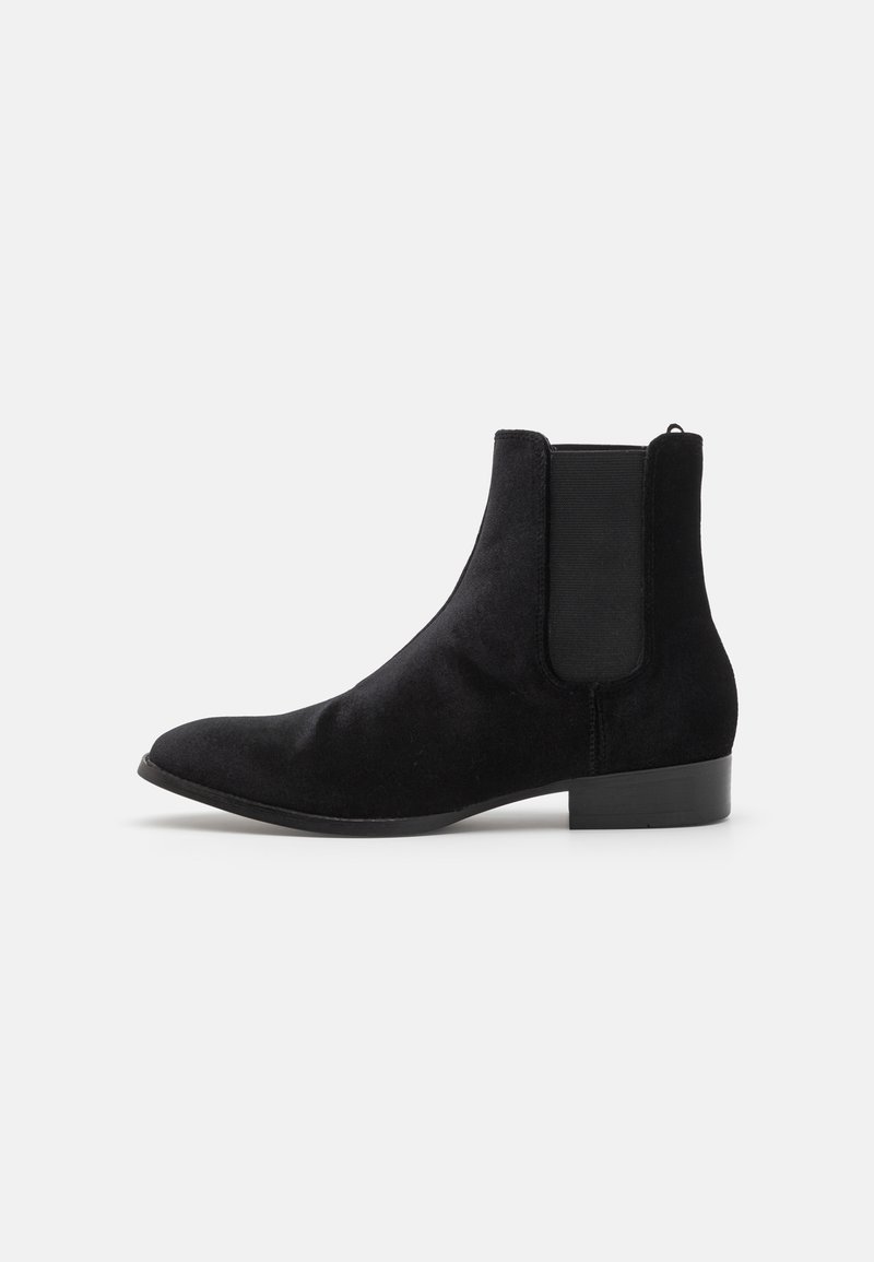 Bottine Chelsea en daim noir avec panneau latéral élastique et petit talon sur fond clair uni.