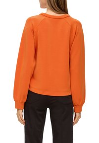 Oranje trui met raglanmouwen en een relaxed fit. Gladde textuur, ronde halslijn en elastische manchetten. Gecombineerd met zwarte broeken.