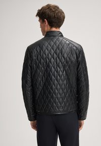 Uomo che indossa una giacca di pelle trapuntata nera e pantaloni scuri, mostrato di spalle su uno sfondo chiaro e semplice.