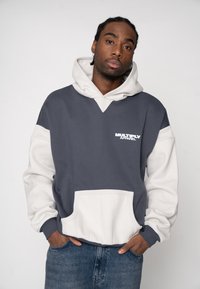 Hanorac cu glugă în gri închis și bej, prevăzut cu buzunar frontal, manșete côtelées și un logo alb pe piept. Textură moale a țesăturii.