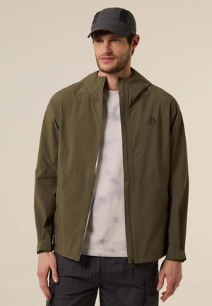 Homme portant une veste à capuche vert olive, un t-shirt blanc tie-dye, un pantalon noir et une casquette noire, se tenant devant un fond uni.