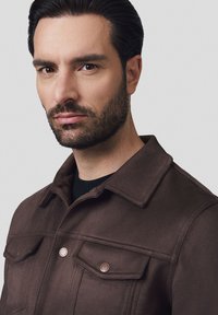 Brązowa wełniana kurtka z klasycznym kołnierzem, dwiema kieszeniami na piersi z guzikami i teksturowanym wykończeniem, noszona na czarnej koszuli.