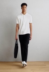 Maglietta polo bianca con maniche corte, pantaloni neri e scarpe bianche con suole nere. Porta una borsa nera ed è in piedi su un pavimento in legno.