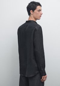Zwarte longsleeve shirt met een zachte textuur, knoopsluiting bij de mouwen en een ronde zoom. Beschikt over een kraag en een detail met een rugnaad.