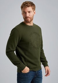 Sweatshirt côtelé vert foncé avec un col rond et un logo circulaire en relief sur la poitrine, associé à un jean en denim.