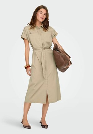 Nő bézs övvel ellátott inget visel, leopárdmintás magassarkú cipőben, nagy, laza barna táskát hord, és vastag karkötőket visel.