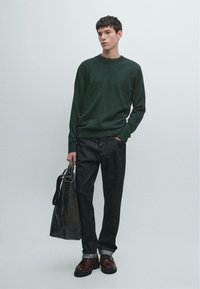 Maglione lavorato a maglia verde scuro, jeans in denim nero, scarpe marroni con lacci. Modello tiene una borsa nera con texture. Design semplice e minimalista.