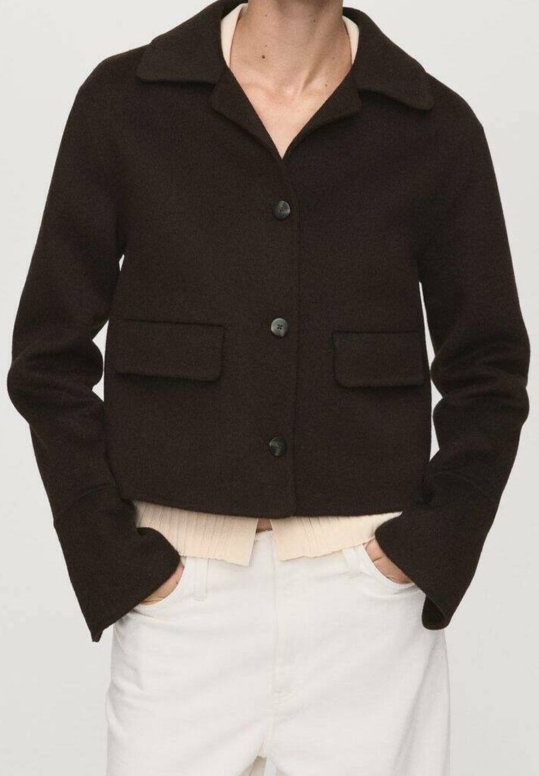 Personne portant une veste boutonnée marron foncé par-dessus un pull clair, avec un pantalon blanc, mains dans les poches, montrée des épaules aux hanches.